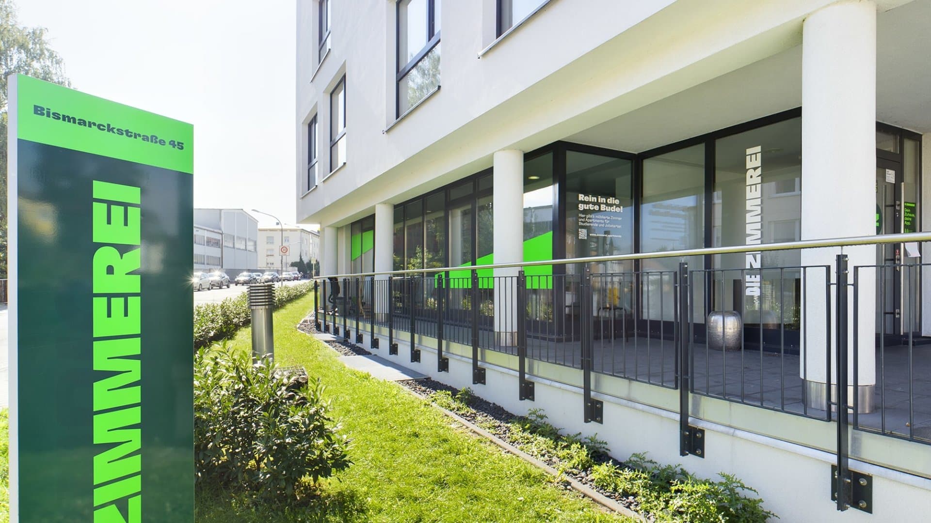 Eingang und Fassade eines modernen Wohngebäudes mit Balkonen und grünem Schild 'Die Zimmerei' an der Bismarckstraße 45