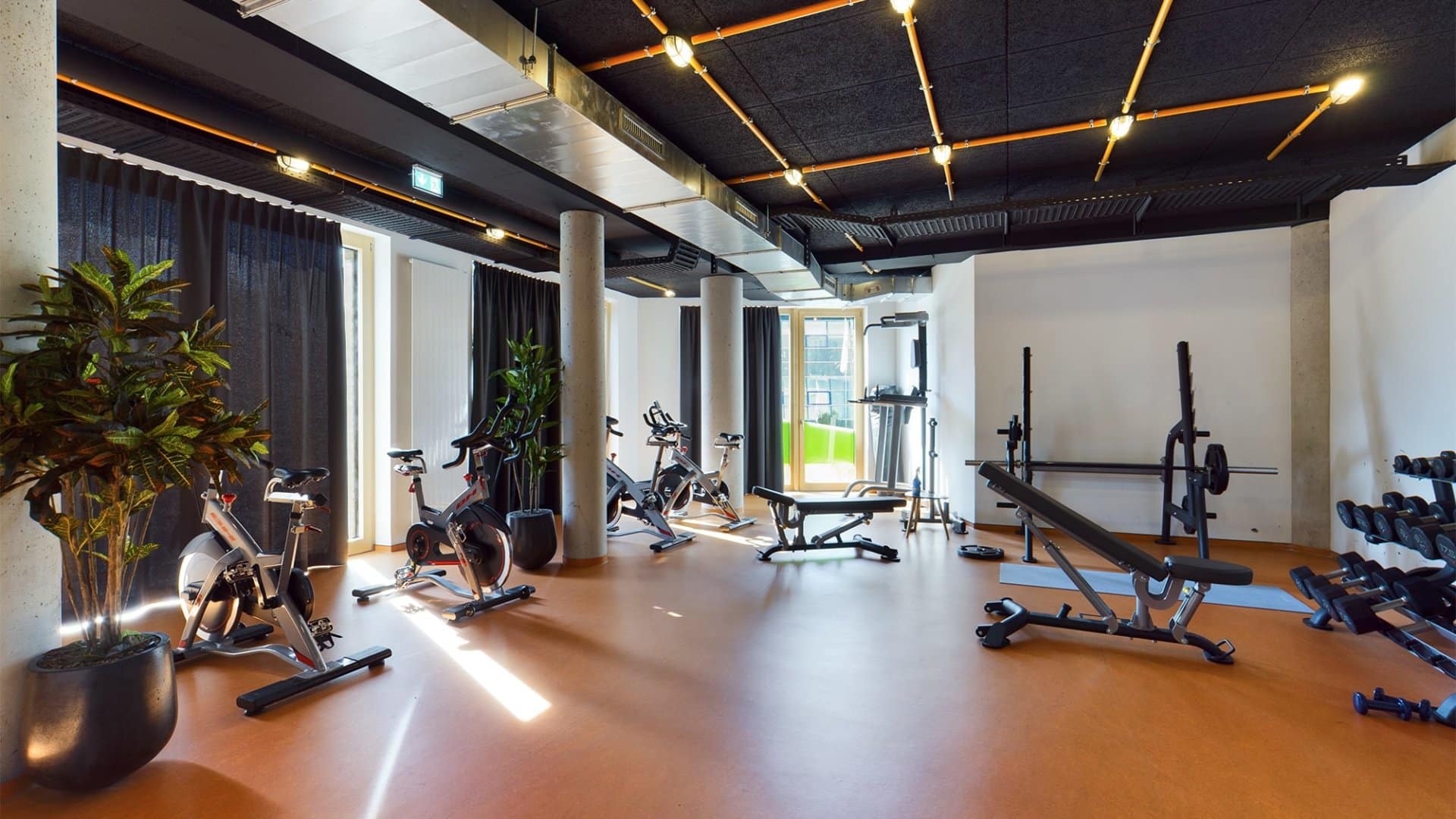 Moderner Fitnessraum mit mehreren Fahrradergometern, Hantelbänken und Krafttrainingsgeräten, große Fenster mit Vorhängen