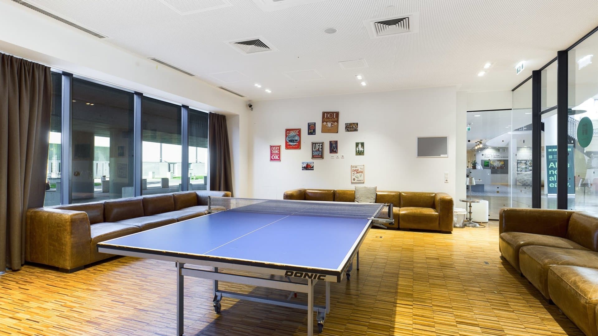 Innenraum mit blauem Tischtennistisch in der Mitte, braunen Ledersofas entlang der Wände und Fenster mit dunklen Vorhängen