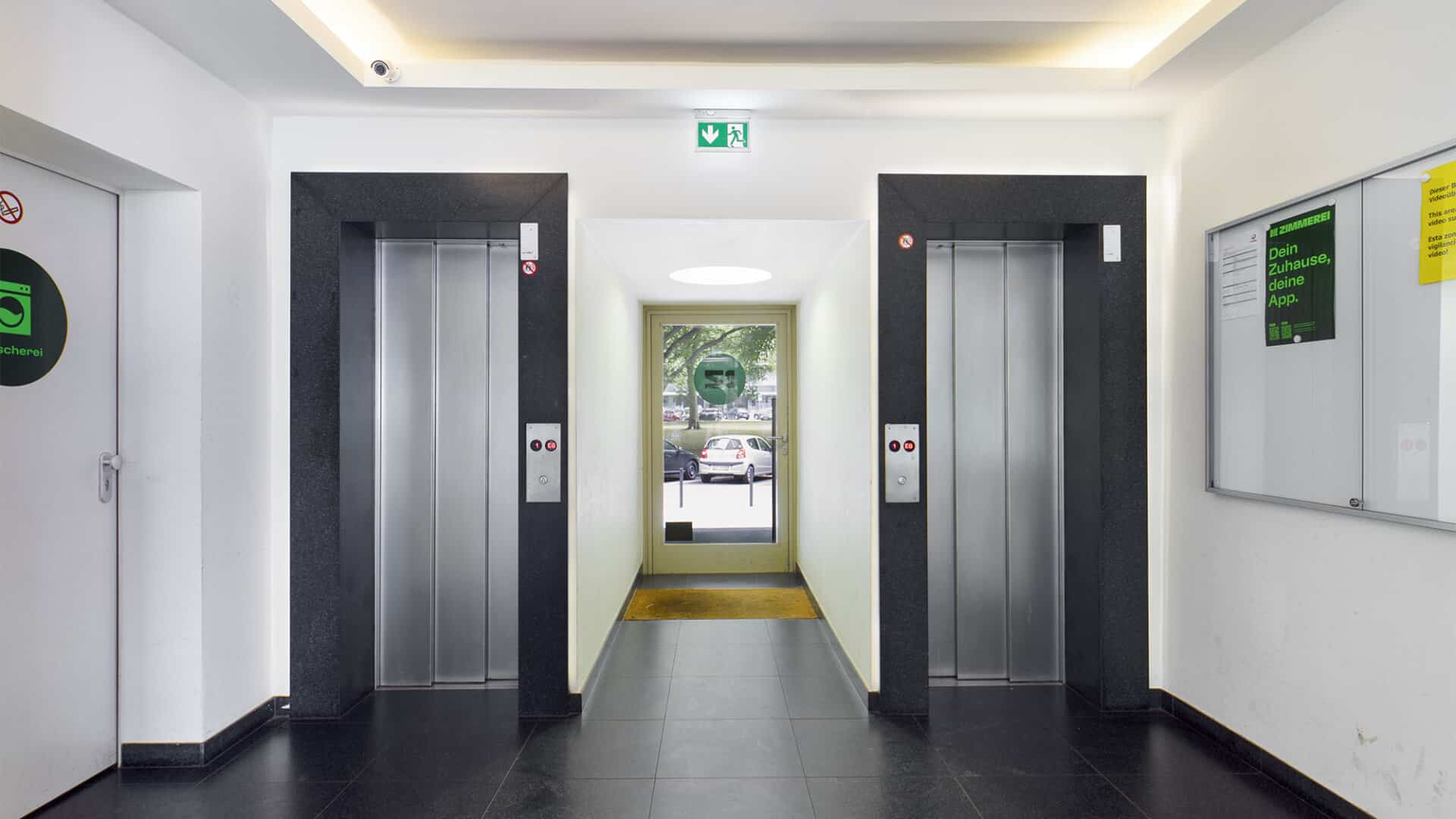Heller Aufzugsvorraum mit zwei Fahrstühlen, schwarzem Fliesenboden und Blick zur Eingangstür.
