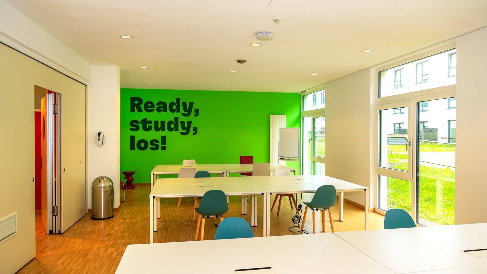 Moderner Arbeitsraum mit weißen Tischen, blauen und weißen Stühlen, grüne Wand mit Text 'Ready, study, los!' und Holzboden