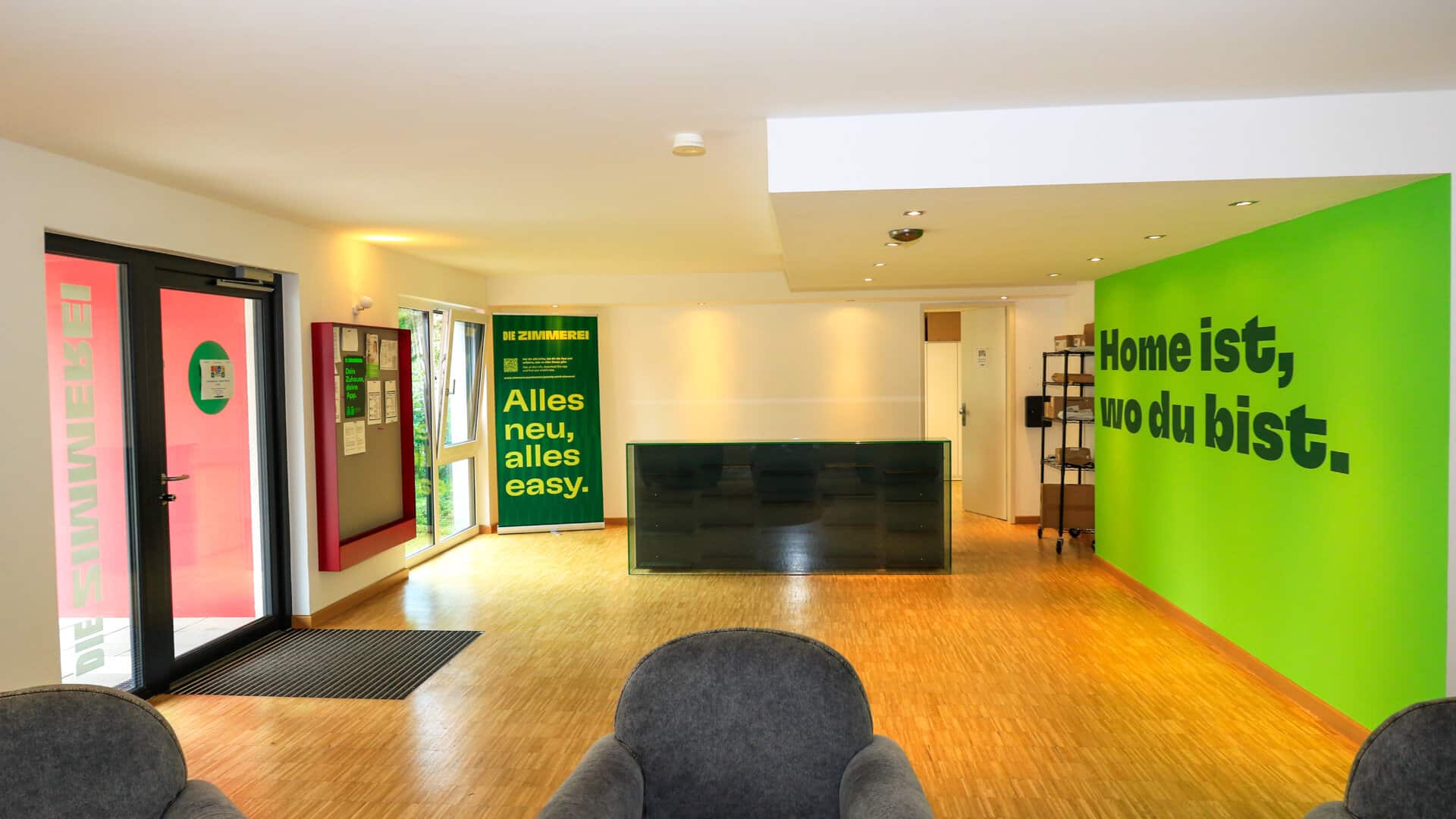 Empfangsbereich mit grüner Wand und Schrift 'Home ist, wo du bist.', schwarzer Empfangstresen und zwei grauen Sesseln