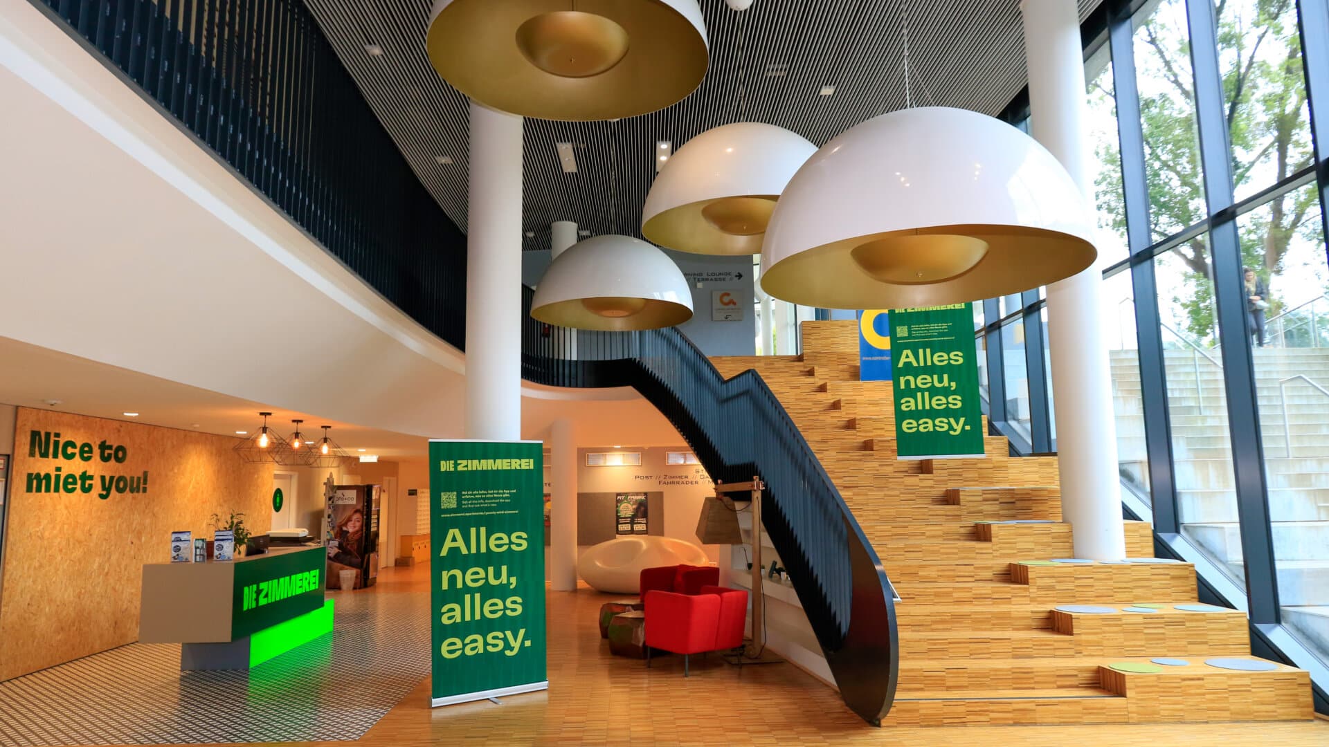 Innenansicht eines modernen Empfangsbereichs mit Treppe, großen Hängeleuchten und Bannern mit dem Text 'Alles neu, alles easy'