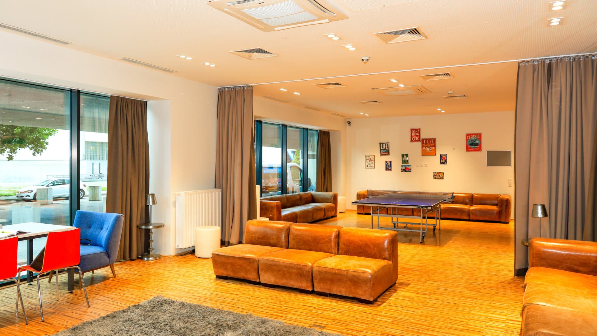 Großer Gemeinschaftsraum mit braunen Ledersofas, Tischtennisplatte, blauem Sessel und Esstisch mit roten Stühlen