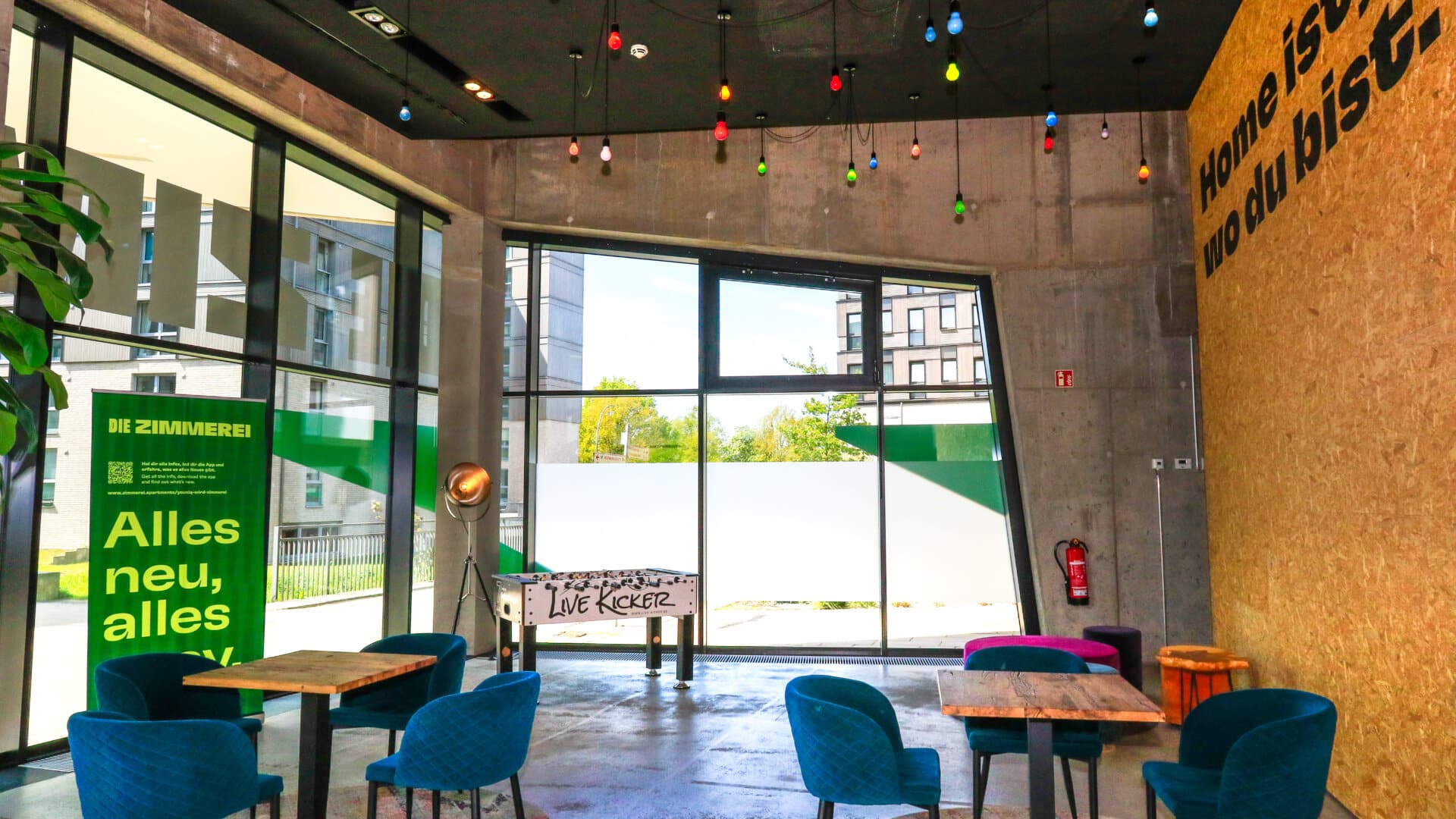 Innenraum eines Cafés mit blauen Stühlen, Holztischen und großen Fenstern, Schriftzug 'Home ist, wo du bist' an der Wand