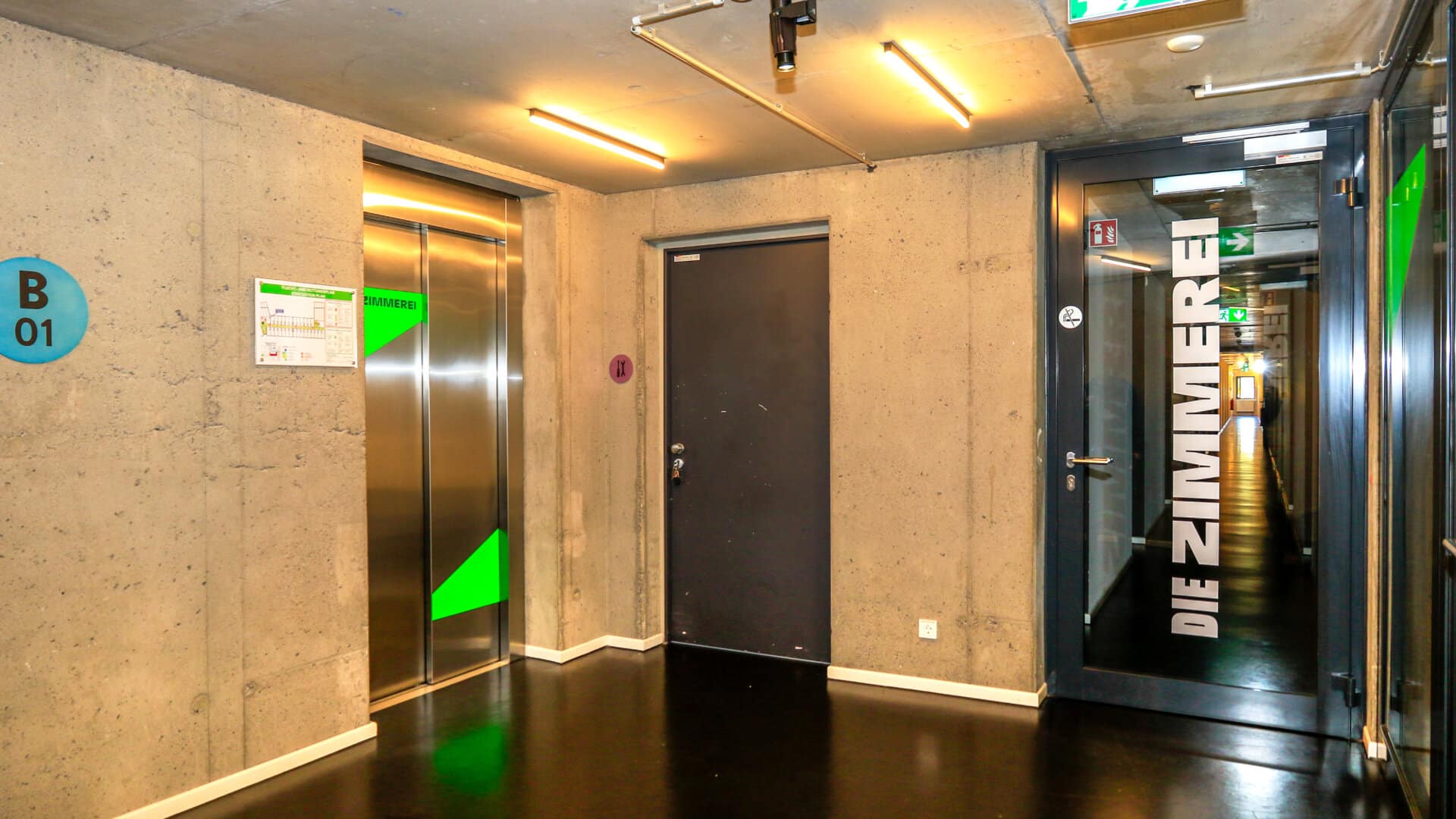 Flur mit Aufzug, schwarzer Tür und Glastür mit Aufschrift 'DIE ZIMMER', Notausgangsschild und Betonwänden