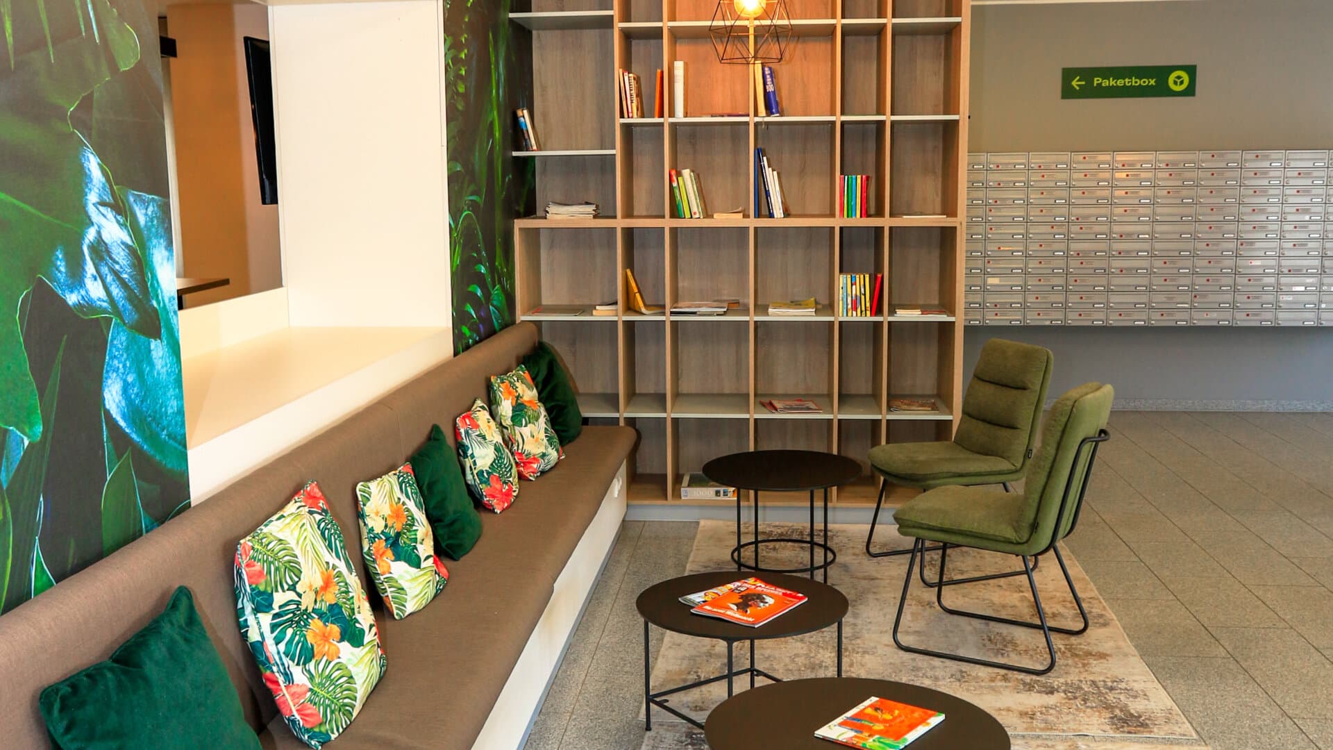 Sitzbereich mit brauner Bank, grünen Kissen, drei runden Tischen, grünen Sesseln, Bücherregal und Briefkästen im Hintergrund