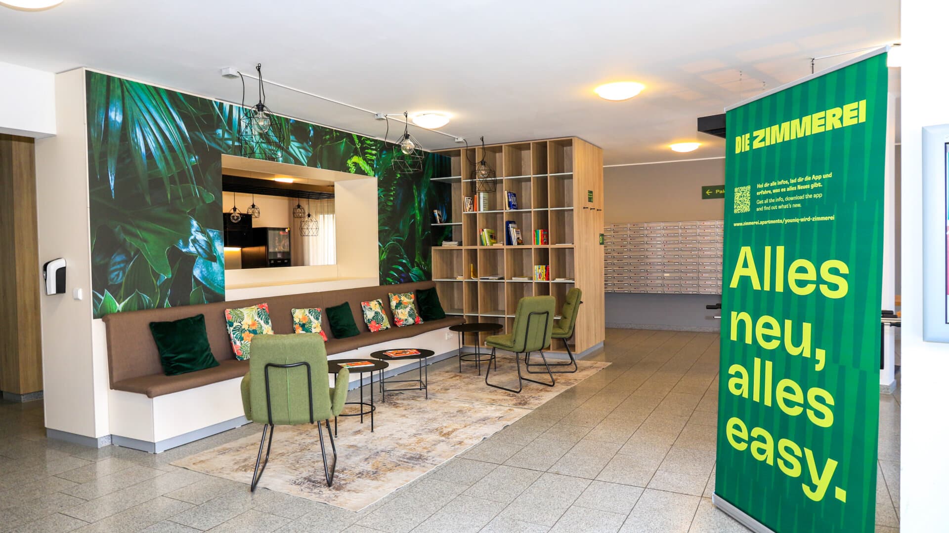 Moderne Lobby mit Sitzbank, grünen Stühlen, großem Spiegel und grünem Banner mit Text 'Die Zimmerei Alles neu, alles easy'