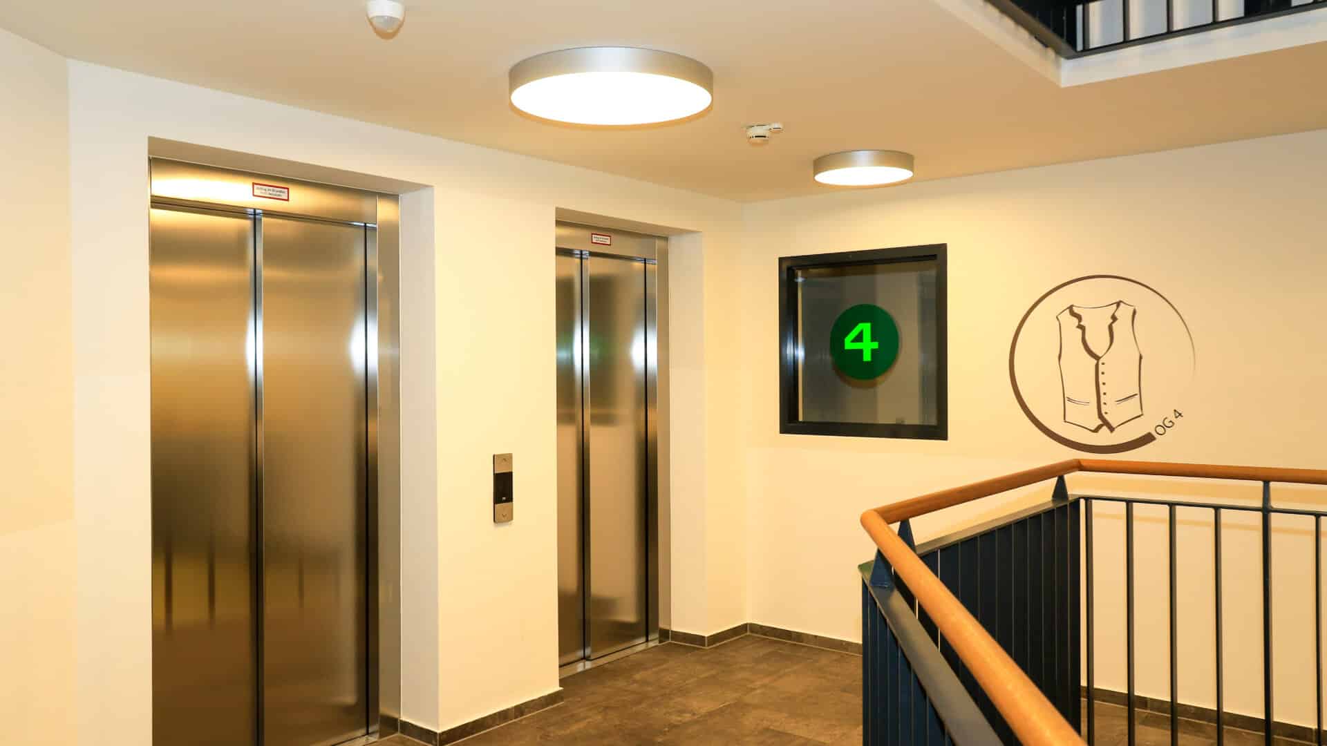 Fahrstühle in einem hellen Treppenhaus mit Anzeige der Etage 4 und einem Wandbild einer Trachtenweste