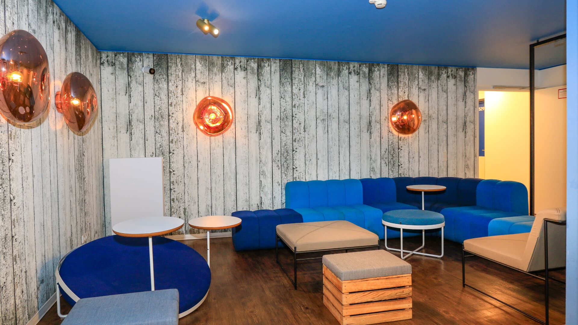 Lounge mit blauen und beigen Sitzmöbeln, Holzboden und Wand mit Holzpaneelen, drei runde Wandlampen
