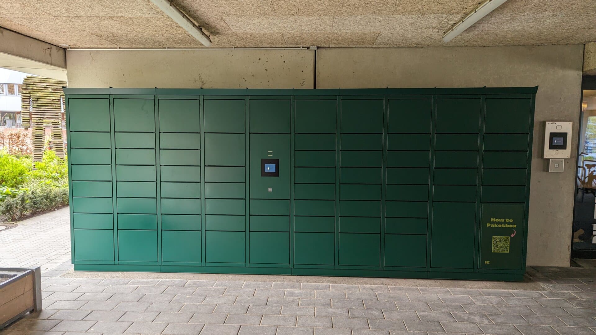Grüne Paketstation mit mehreren Fächern und digitalem Display an einer Wand unter einem Betonüberhang